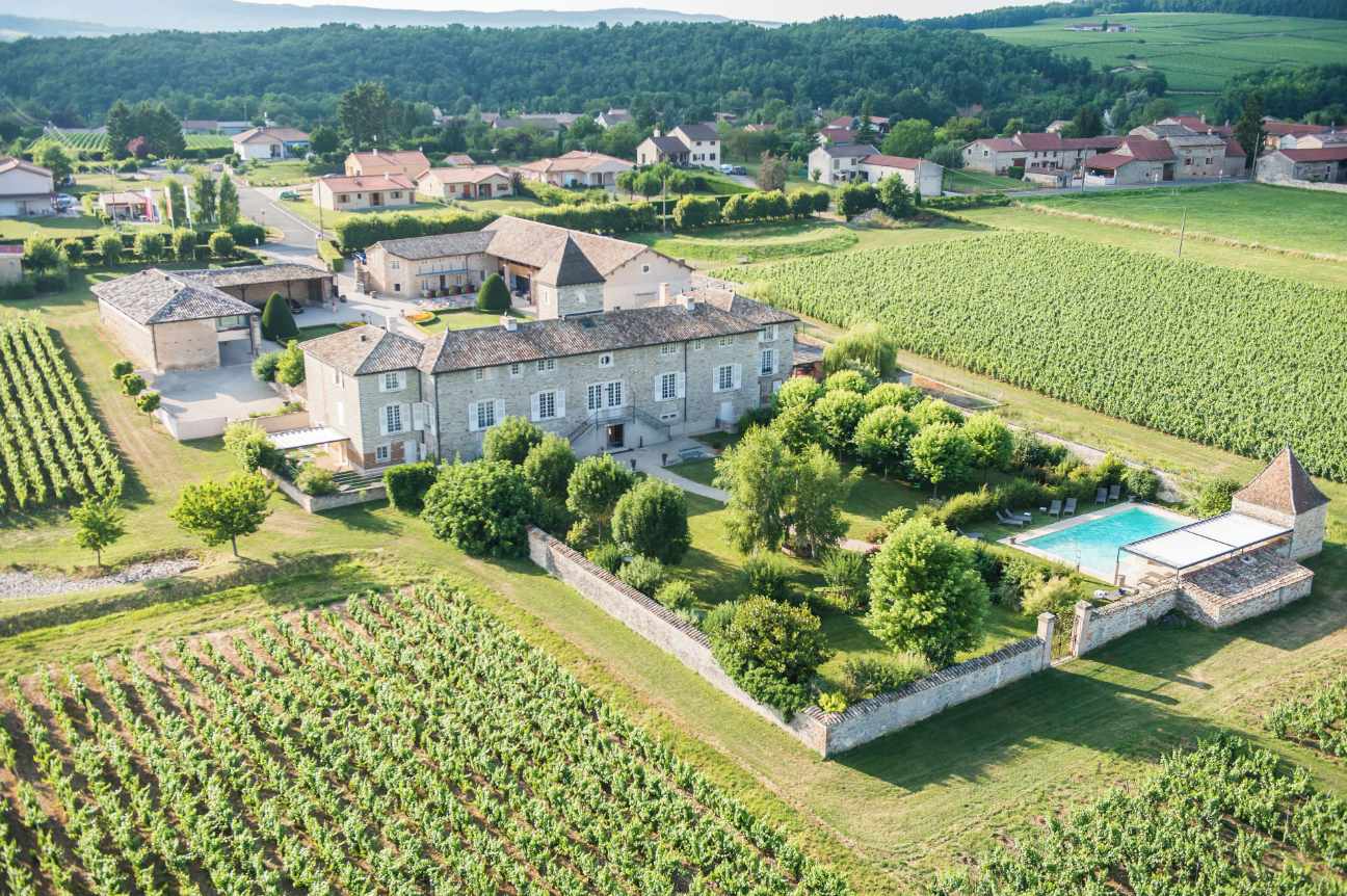 Aerial view of a stone château and its outbuildings enclosed by walls, with vineyard rows running up to the property on two sides